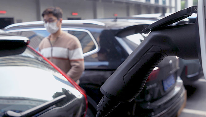 Close-up of a smart EV charging plug with enhanced safety features, with a person wearing a mask standing near cars in the background.
