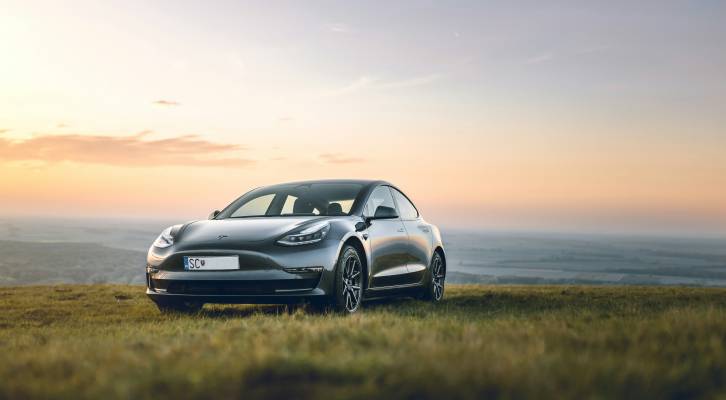 Silver electric car parked on a grassy hilltop at sunset, overlooking a vast landscape.