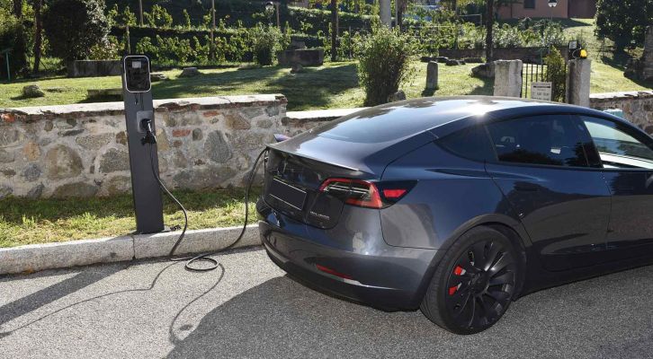 A dark gray electric car charging at a BESEN BS20 7kW EV charging station, located in a serene park-like area with stone walls and greenery.