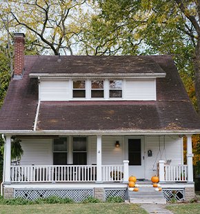 Front view of a white single-family home with a porch and pumpkins on the steps, surrounded by trees.