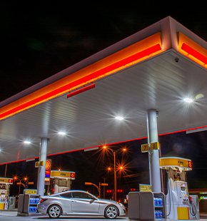 Gas station at night with a white car parked under the illuminated canopy, surrounded by fuel pumps.
