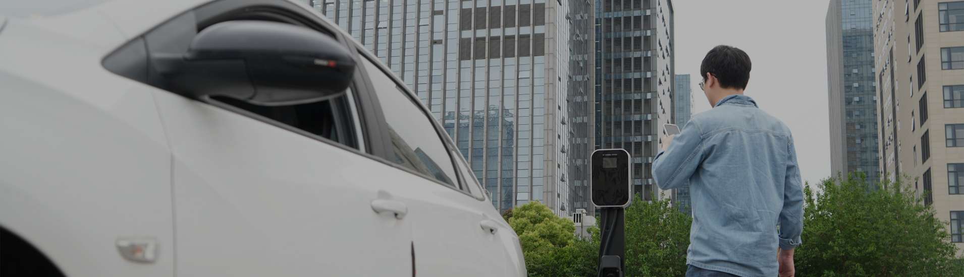 A person standing near an app electric vehicle charger, with a white car parked nearby and modern buildings in the background.