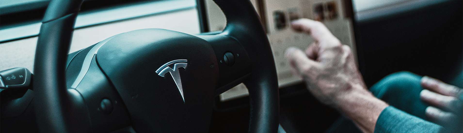 Interior of a Tesla Model 3 with a focus on the steering wheel displaying the Tesla logo, and a driver's hand interacting with the touchscreen control panel