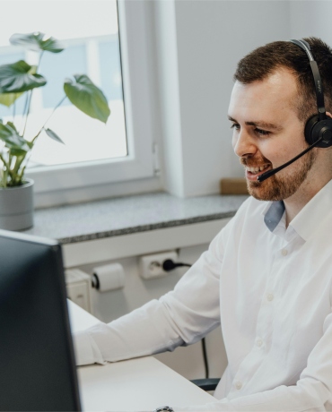 A friendly male customer service representative wearing a headset, smiling as he assists a customer, seated at a white desk in a bright office with a window and green plants in the background.