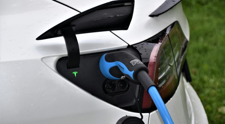 Close-up of a white Tesla electric car being charged with a blue Type 2 connector plugged into the rear charging port, showing the illuminated Tesla logo and tail light.