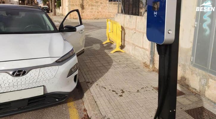 Parked electric vehicle beside a pole-mounted curbside EV charger on a city sidewalk, with a play button indicating a video.