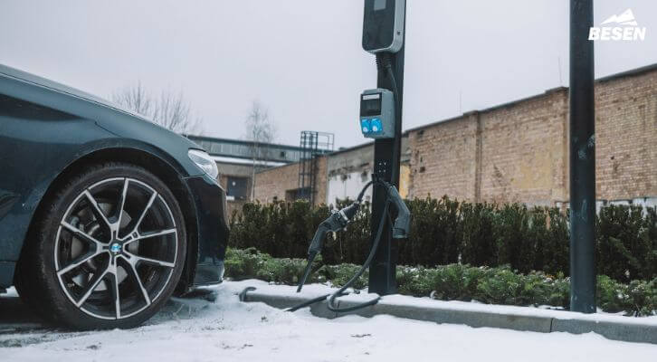 Snow-covered curbside electric vehicle charger with a black car parked beside it, shown with a play button overlay for video content.