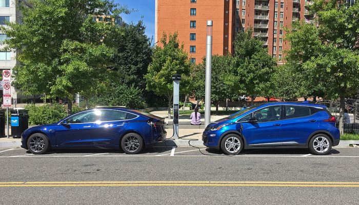 A Tesla Model 3 and a Chevrolet Bolt EV, both blue, parked side by side while charging at a public EV charging station on a city street.