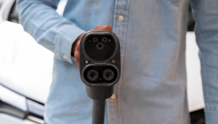 Person in denim shirt holding a CCS electric vehicle charging plug close to the camera, showing the dual-pin DC fast-charging connectors.