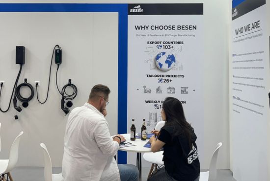 Two people seated at a table in a BESEN exhibition booth discussing EV charging products, with wall-mounted chargers and marketing posters in the background.